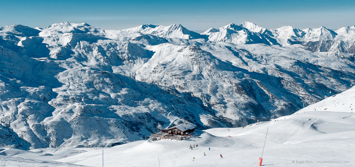 High, wide view of skiers heading for mountain restaurant in Les menuires ski resort with mountains in background