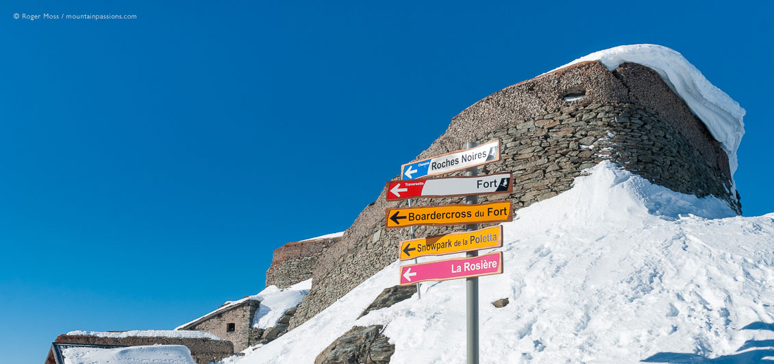 Low, wide view of ski piste sign with sow-covered fort above La Rosiere