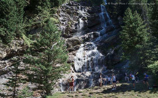 Walkers beside rockface among trees with waterfall