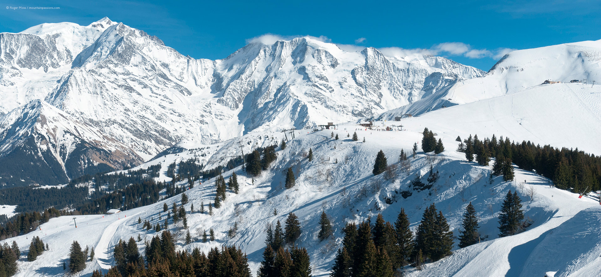 Wide view of snow-covered mountains, with distant ski lift, pistes and Mont-Blanc in background.