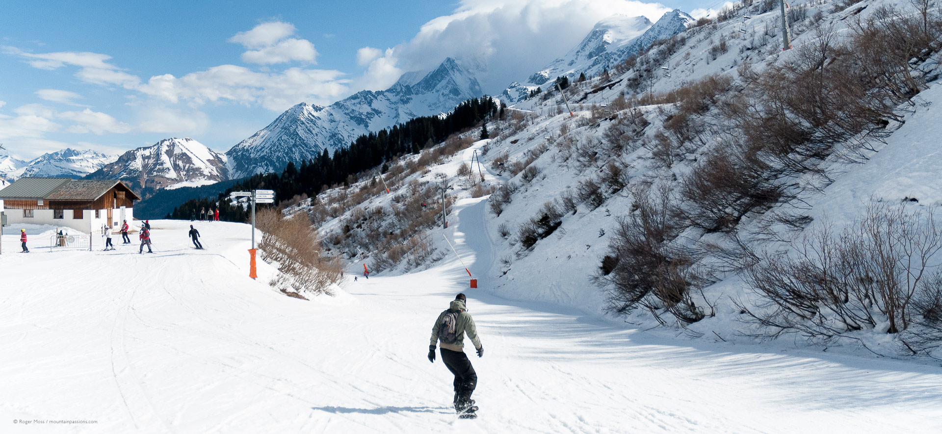 Snowboarder and skiers at junction of pistes with mountain background.