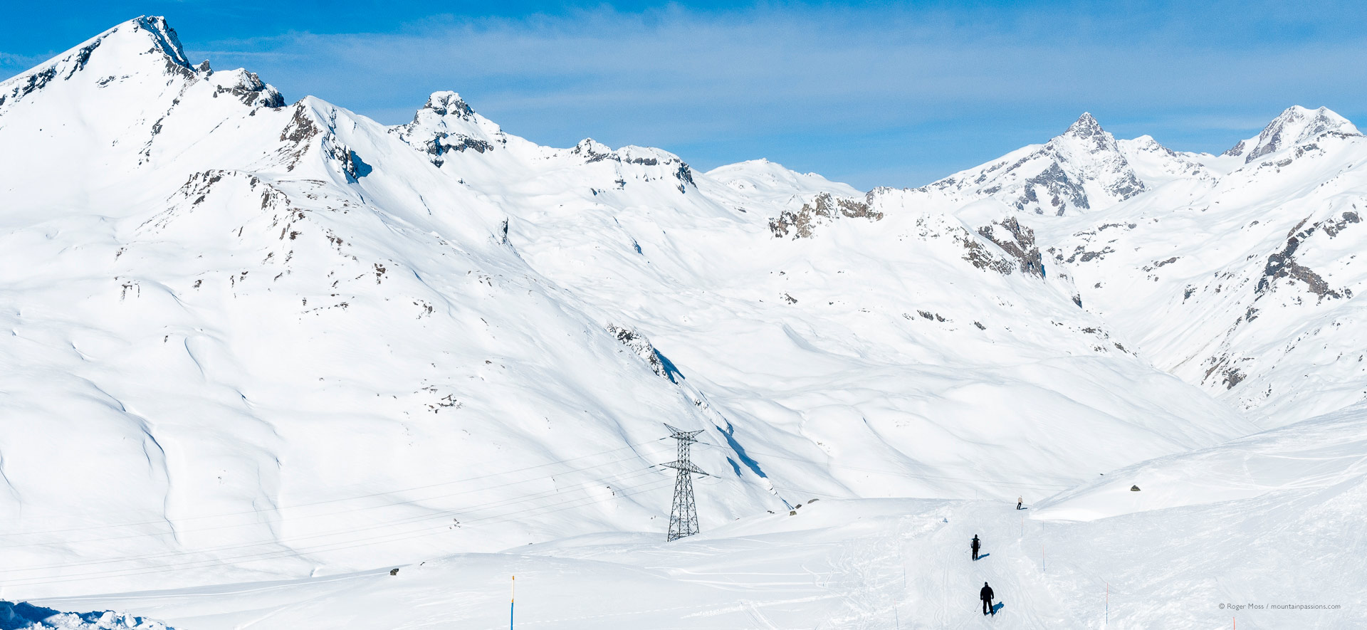 Rear view of two skiers on piste among vast snowy mountain landscape.