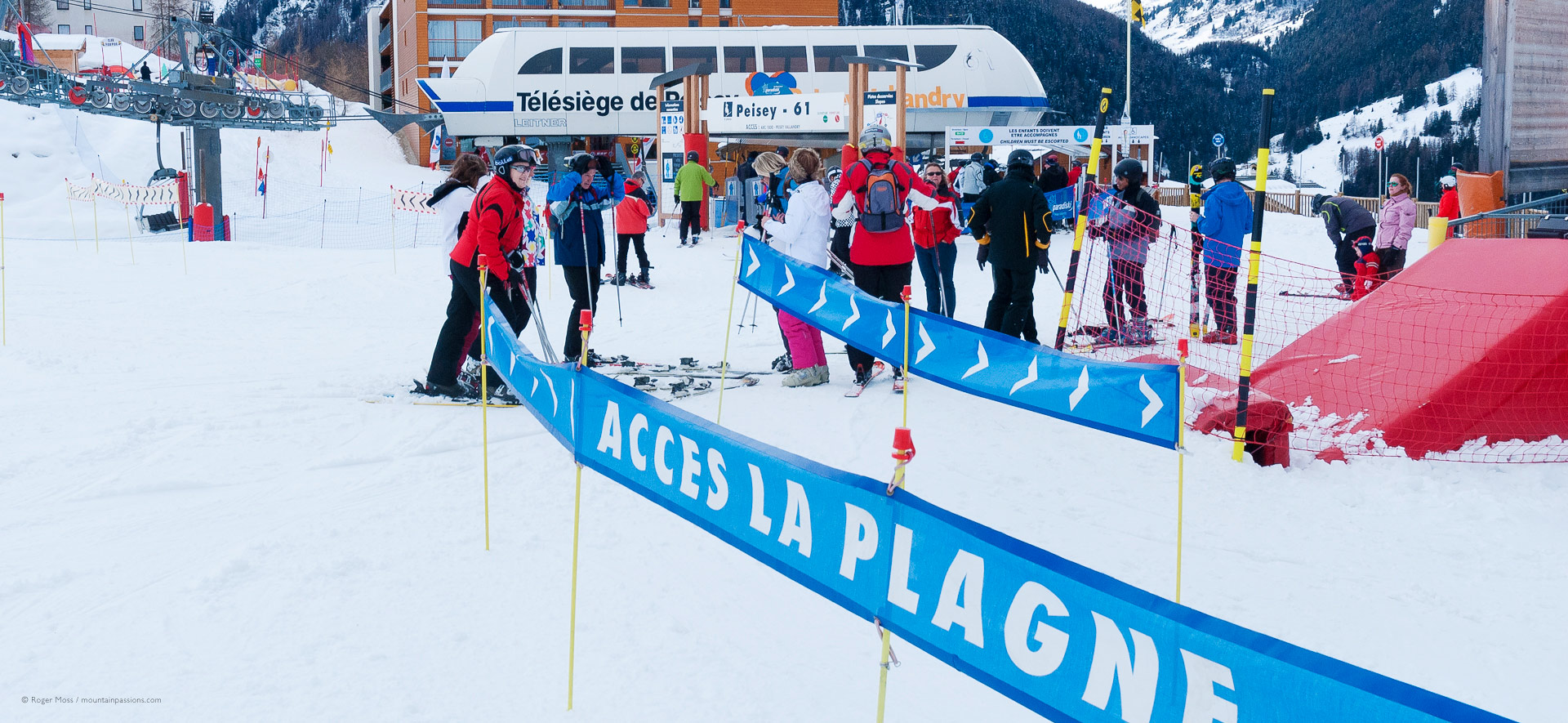 Skiers beside signs to liason with La Plagne, with ski-lifts in background.