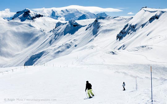 Rear view of two skiers descending wide piste with big mountain view