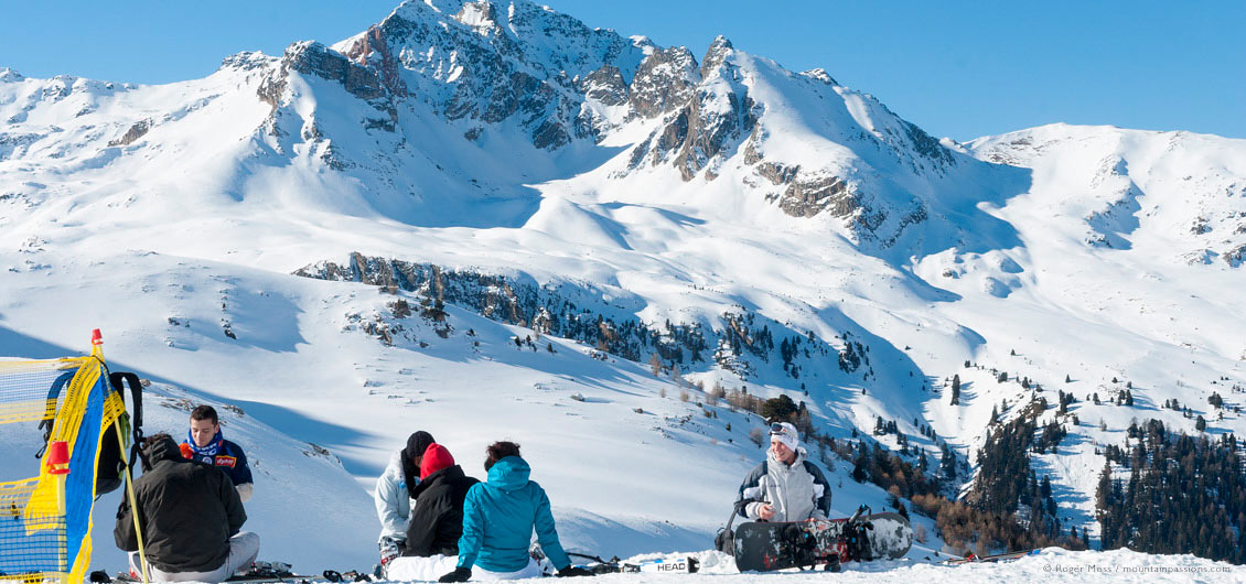 Young skiers and snowboarders picnic beside piste, with big mountain backdrop