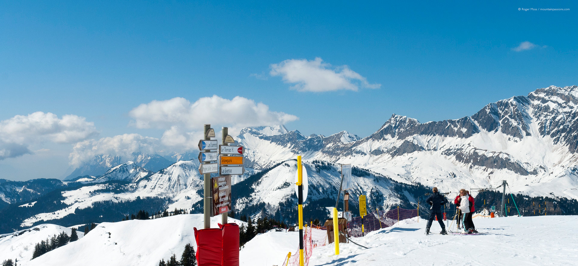 Wide view of skiers beside piste signage with mountains at Combloux.