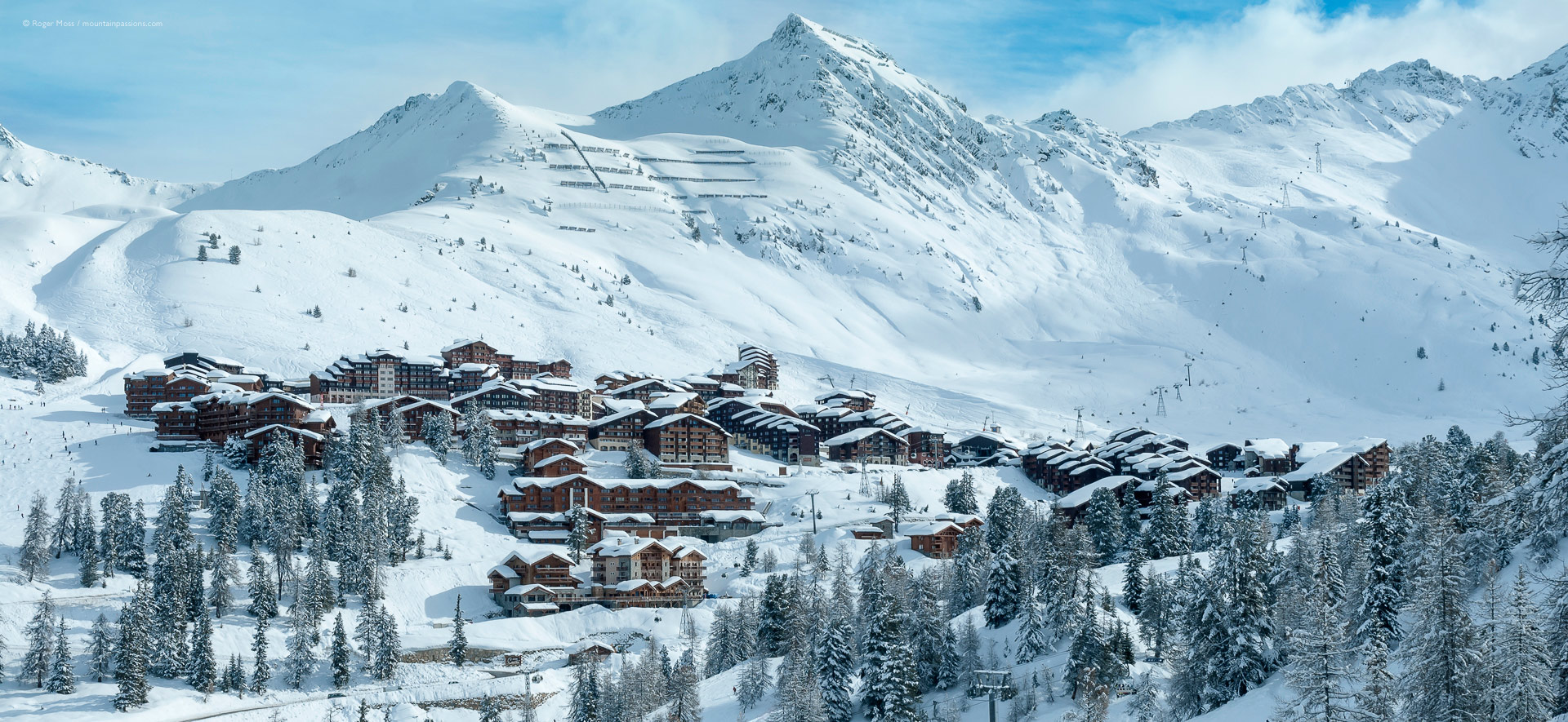 General view of ski village and mountains beyond snow-covered trees.