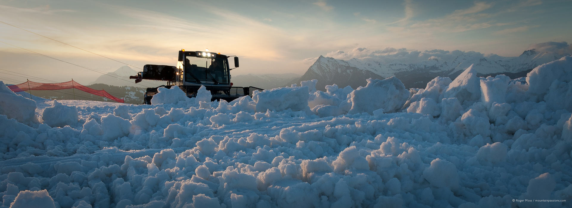 Low view of piste groomer at dusk, beyond banked-up snow