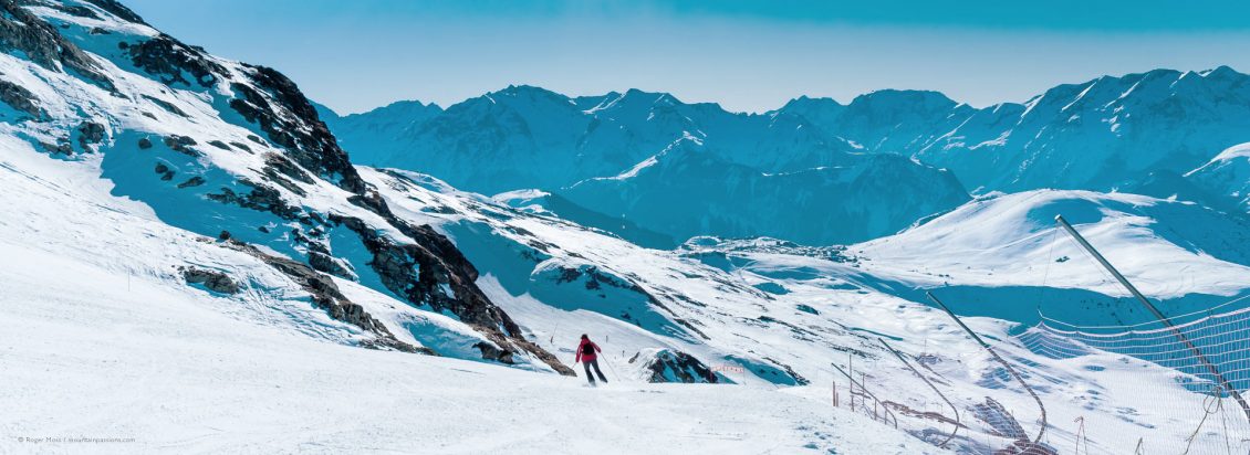 Wide view of skier on piste with mountains and Alped'Huez ski village in background.