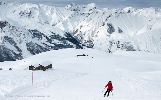 Rear overview of skier passing chalets, with big mountain background