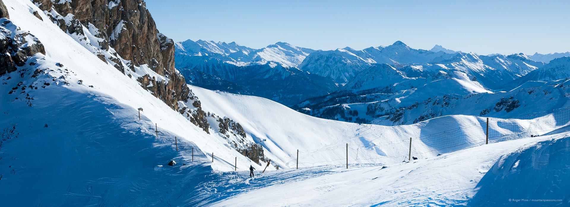 Long view of lone skier on snow-covered ridge with vast mountain background