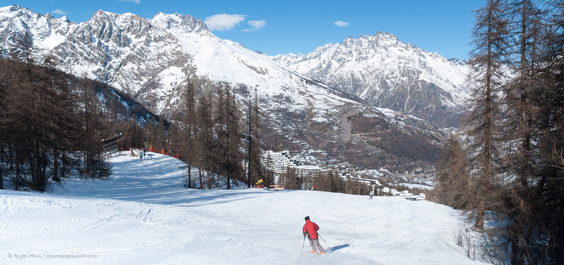 Skier on wide, tree-lined piste, with Puy Saint Vincent ski village below