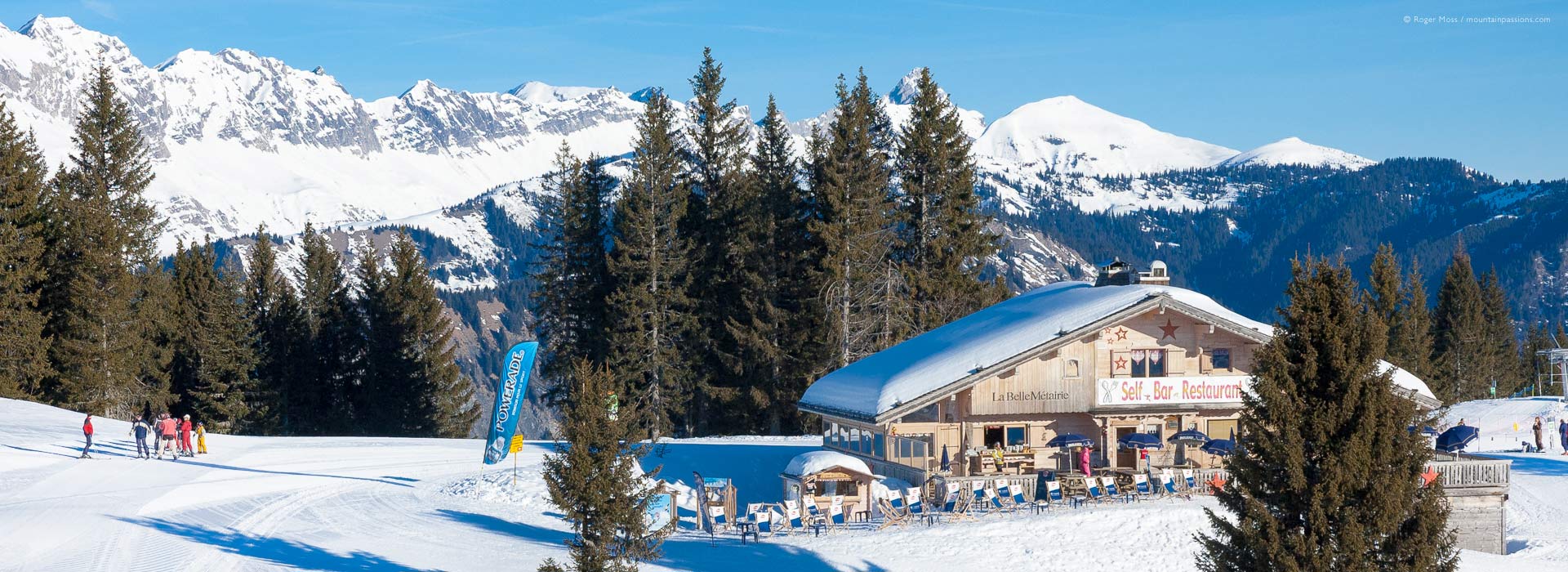 Wide view of early morning skiers beside chalet-style mountain restaurant above Crest Voland.