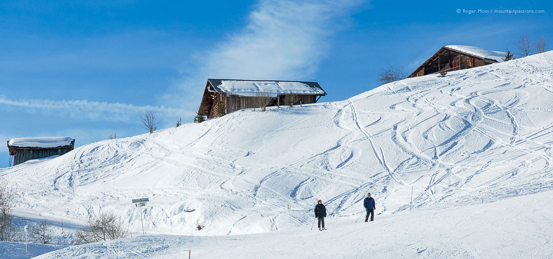 Low, wide view of two skiers with mountain chalets near Crest Voland ski resort