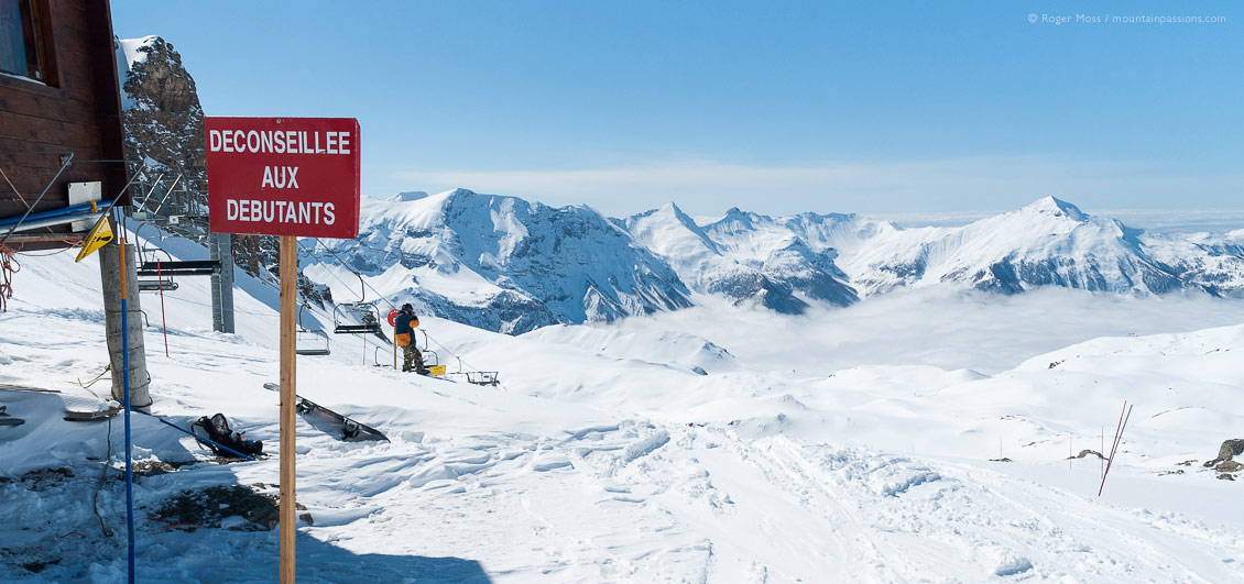 Wide view past piste signage to start of ski run, with snowboarder at Orcieres 1850