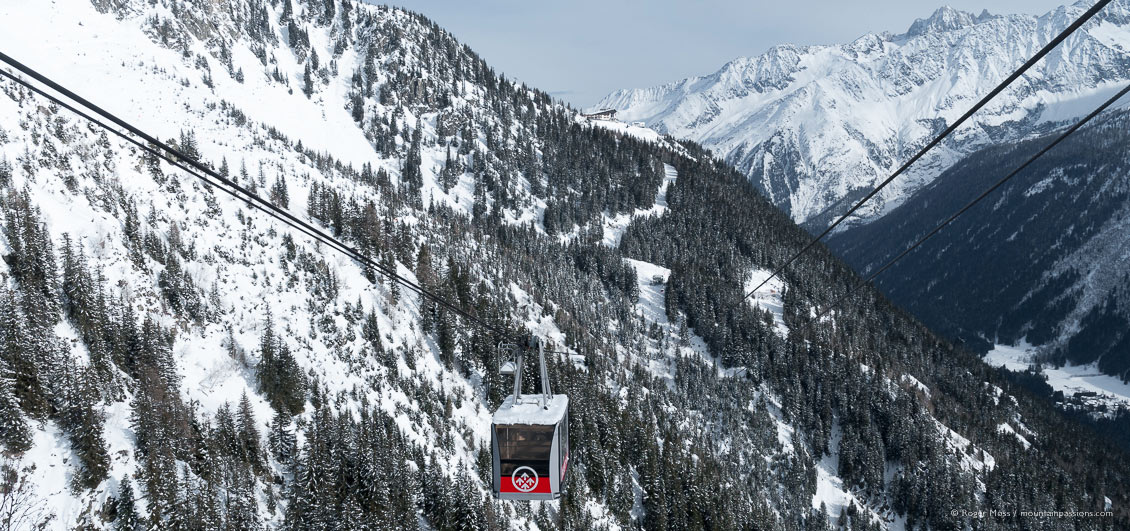 Wide view of cable-car crossing snow-covered valley, showing ski pistes and mountains.