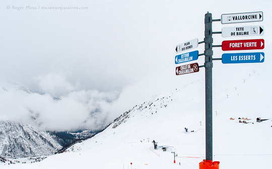 View past ski piste signage to snow-covered valley with clouds building