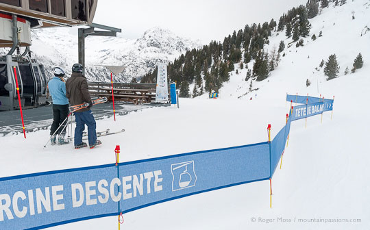 Wide view of two skiers beside top station of gondola ski lift, with piste signage above Vallorcine