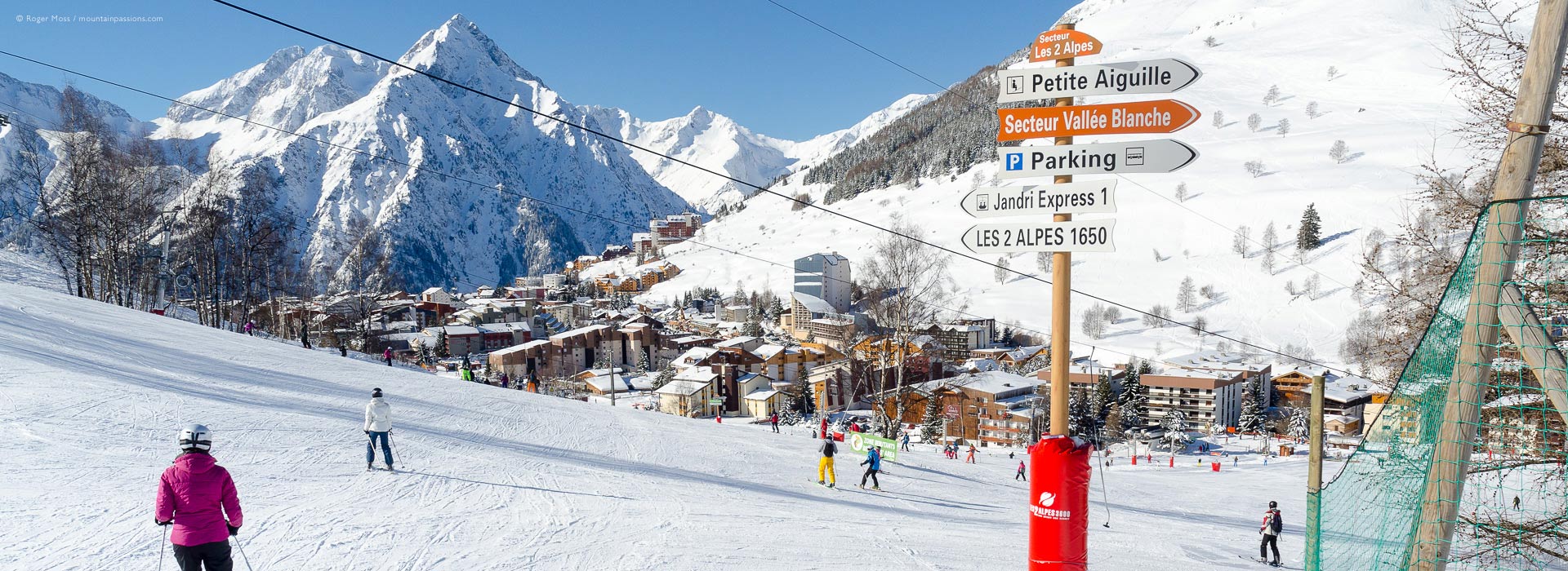 Wide view of skiers approaching ski lifts, with Les 2 Alpes ski village and mountains